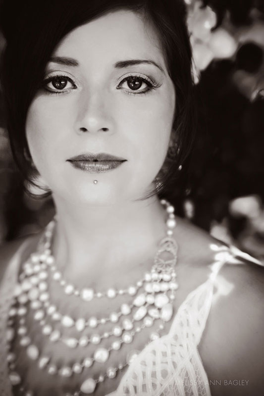 Black and white head and shoulders fine art portrait of a woman with dark hair and a piercing under her lips. She is wearing a multi strand necklace and white dress. She is looking directly at the camera.