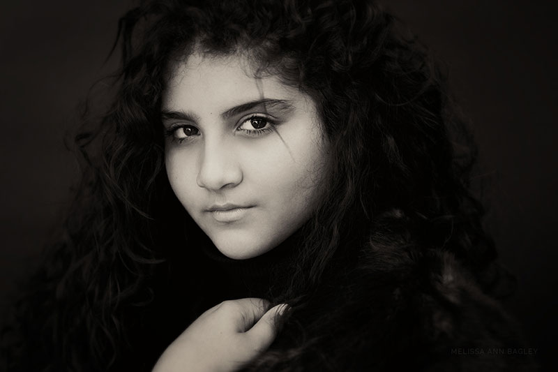 Black and white fine art portrait of a young woman with curly long dark hair against a dark backdrop. She is looking over her shoulder at the camera and one of her hands is holding the lapel of her coat.