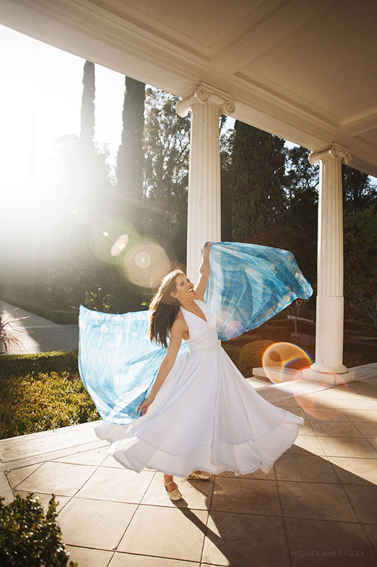 Color fine art portrait of a female Persian dancer in white dress with a blue veil spinning while back lit by the sun. She has stone pillars and plants behind her.