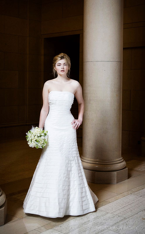 Bridal fashion photograph of a blond model standing in front of a stone pillar and holding a bouquet of white flowers.