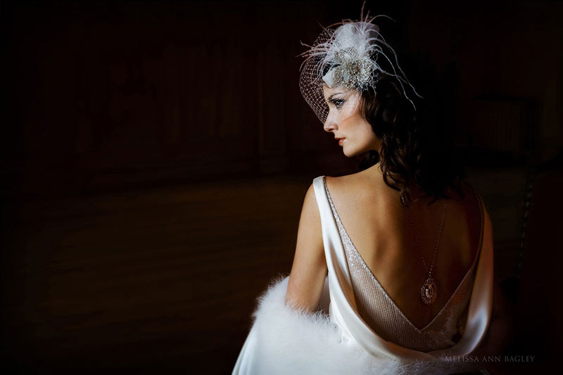 Color bridal fashion photograph of a model wearing a wedding dress with a deep v in the back that is framing a long necklace. She is seated with her back to the camera and is looking over her left shoulder in profile.