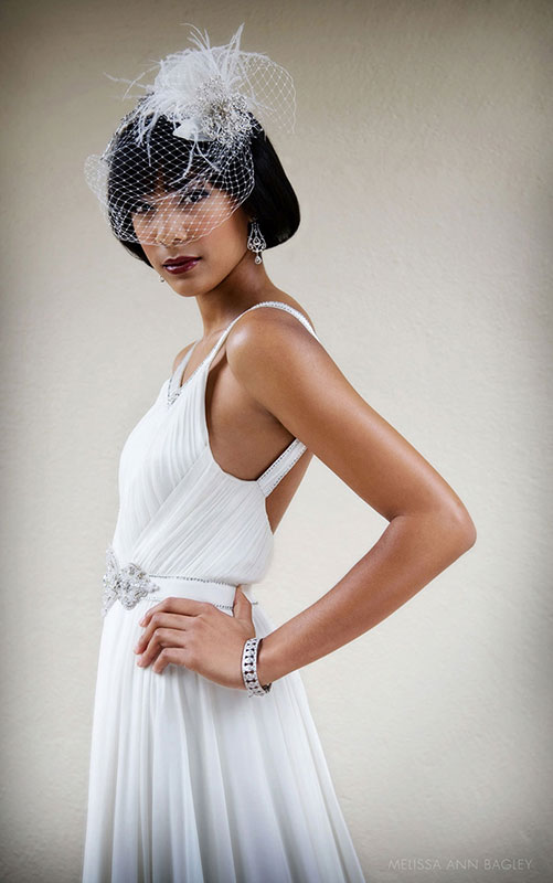 Bridal fashion photograph of a dark haired model in a wedding dress and hair accessory. She is turned away from the camera and looking back over her shoulder with her hand at her hip.