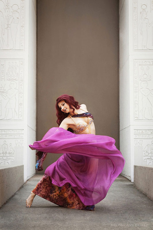 Color fine art portrait of a woman with long red hair in a purple and gold belly dance costume. The walls behind her are cream and have Egyptian hieroglyphics on them.