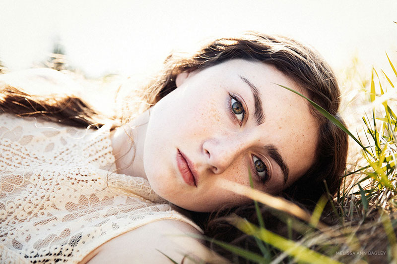 Color fine art portrait of a young woman with long dark hair and brown eyes laying in a grassy field. She is wearing a cream colored lace top and looking directly at the camera.