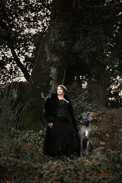 Color fine art portrait of a woman with short hair wearing a black steampunk dress and coat standing in a forest with her large grey dog on a leash.