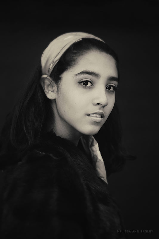 Black and white fine art portrait of a young woman with shoulder length hair. Her body is turned away from the camera and her head is turned toward the camera. She is wearing a dark coat and has a light colored scarf in her hair.