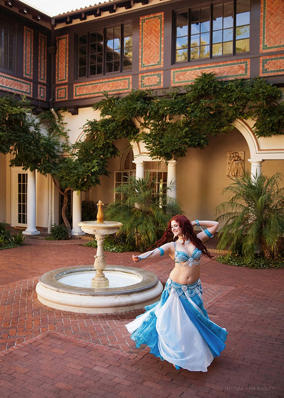 Color fine art portrait of a woman with long red hair in a blue and white belly dance costume twirling in an outdoor courtyard.
