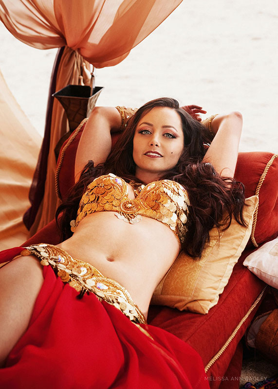 Color fine art portrait of a woman with long dark hair in a red and gold belly dance costume. She is laying back on a red lounging couch.