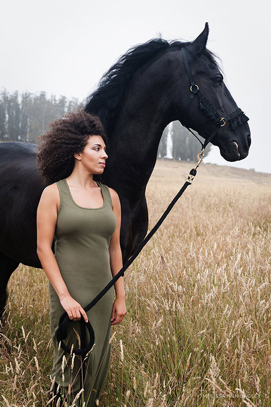Color fine art portrait of a woman in a green tank dress standing in a field next to a large black horse. Both are shown in profile.