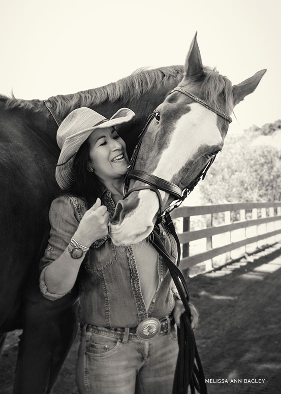 Black and white fine art portrait of a woman wearing a denim top, jeans and cowboy hat with her horse standing behind her. The horse is turning his head to touch her hand.