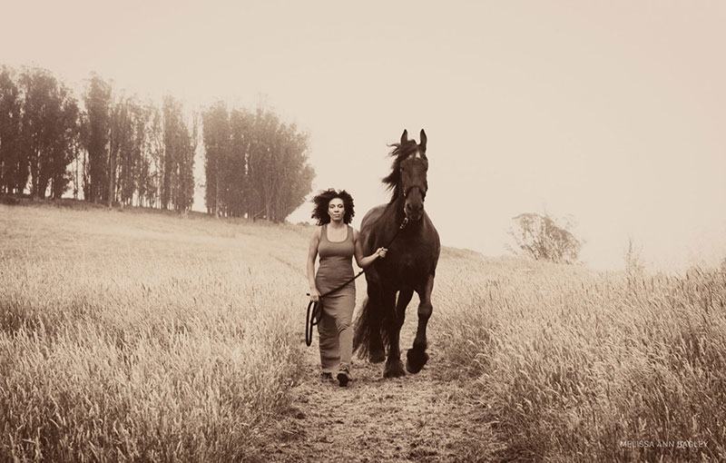 Sepia colored fine art portrait of a woman in a tank dress walking a large horse through an open field with trees in the background.