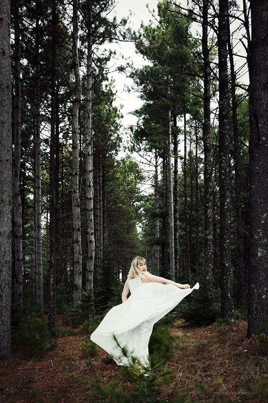 Color fine art portrait of a blond haired woman in a white dress. She is standing in a forest surrounded by trees and is holding the hem of her dress.